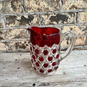 May include: A clear glass pitcher with a ruby red top and a decorative pattern of red hexagons. The pitcher has a clear handle and a spout for pouring. The pitcher is sitting on a wooden surface.