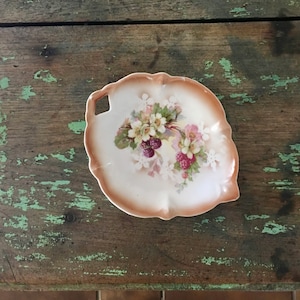 May include: A white porcelain leaf-shaped dish with a floral design featuring pink and white flowers and dark purple berries. The dish has a brown rim and a small handle.