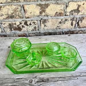 May include: A bright green glass vanity set featuring a rectangular tray with a sunburst pattern, a lidded jar, a smaller lidded container, and a ring holder. The pieces are made of translucent green glass with faceted edges and geometric detailing.