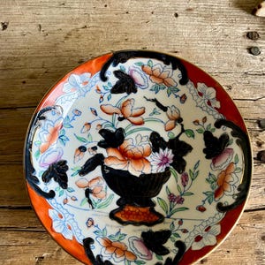 May include: A white porcelain saucer with a black and orange floral design. The saucer has a red rim and a gold trim.
