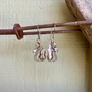 May include: A pair of silver earrings with a leaf design. Each earring features a small, round, light pink gemstone and a smaller, yellow gemstone. The earrings are suspended from a hook and are displayed on a rusty metal rod.