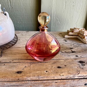 Puede incluir: Un frasco de perfume vintage de vidrio en forma de l&aacute;grima, en tonos rosa y &aacute;mbar. El frasco tiene un tap&oacute;n redondo y se encuentra sobre una superficie de madera r&uacute;stica. El vidrio refleja la luz, creando un brillo c&aacute;lido. Una pieza decorativa.