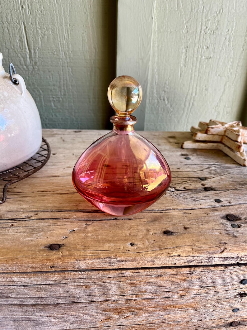 Puede incluir: Un frasco de perfume vintage de vidrio en forma de l&aacute;grima, en tonos rosa y &aacute;mbar. El frasco tiene un tap&oacute;n redondo y se encuentra sobre una superficie de madera r&uacute;stica. El vidrio refleja la luz, creando un brillo c&aacute;lido. Una pieza decorativa.