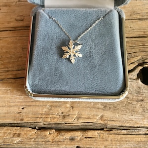 May include: A silver snowflake pendant necklace displayed in a light blue jewelry box. The snowflake pendant is intricately detailed and hangs from a delicate chain. The box has a soft, velvet-like interior and a silver trim.