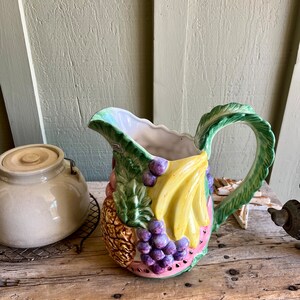 May include: A ceramic pitcher shaped like a watermelon with a handle shaped like a banana leaf. The pitcher is decorated with a pineapple, grapes, and bananas.