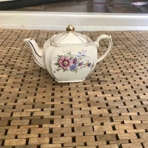May include: A white teapot with a floral design and gold accents. The teapot has a square body and a lid with a gold knob. The teapot is sitting on a wooden surface.