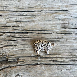 May include: A silver-colored pig-shaped brooch with intricate scrollwork detailing. The brooch is set against a weathered, light brown wooden surface, highlighting its ornate design and metallic sheen.