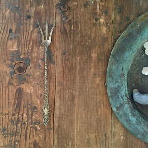 May include: A silver ornate fork with three tines, resting on a wooden surface. The fork has a decorative design and a rounded handle.
