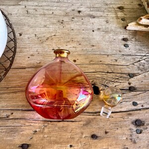 Puede incluir: Una botella de vidrio iridiscente en forma de pera con borde dorado y tap&oacute;n separado. La botella cambia de rojo-naranja a dorado. El tap&oacute;n es transparente con la parte superior dorada. Sobre una superficie de madera envejecida.