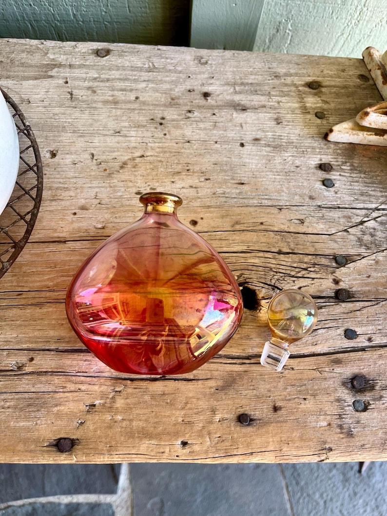 Puede incluir: Una botella de vidrio iridiscente en forma de pera con borde dorado y tap&oacute;n separado. La botella cambia de rojo-naranja a dorado. El tap&oacute;n es transparente con la parte superior dorada. Sobre una superficie de madera envejecida.