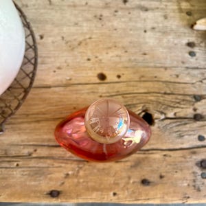Puede incluir: Una botella de perfume de vidrio rosa con un tap&oacute;n redondo y transparente. La botella tiene forma redondeada y est&aacute; sobre una superficie de madera. El tap&oacute;n es de un tono m&aacute;s claro que la botella.