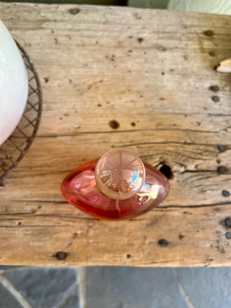 Puede incluir: Una botella de perfume de vidrio rosa con un tap&oacute;n redondo y transparente. La botella tiene forma redondeada y est&aacute; sobre una superficie de madera. El tap&oacute;n es de un tono m&aacute;s claro que la botella.