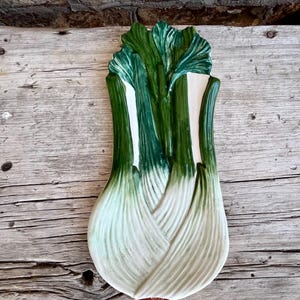 May include: A ceramic decorative plate shaped like a green and white leek. The plate has a detailed design with textured leaves and a realistic appearance. The plate is placed on a weathered wooden surface.