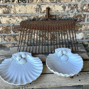 May include: Two white, seashell-shaped decorative dishes with detailed shell and leaf accents. An antique metal rake is positioned above the dishes against a weathered brick wall and wooden surface.