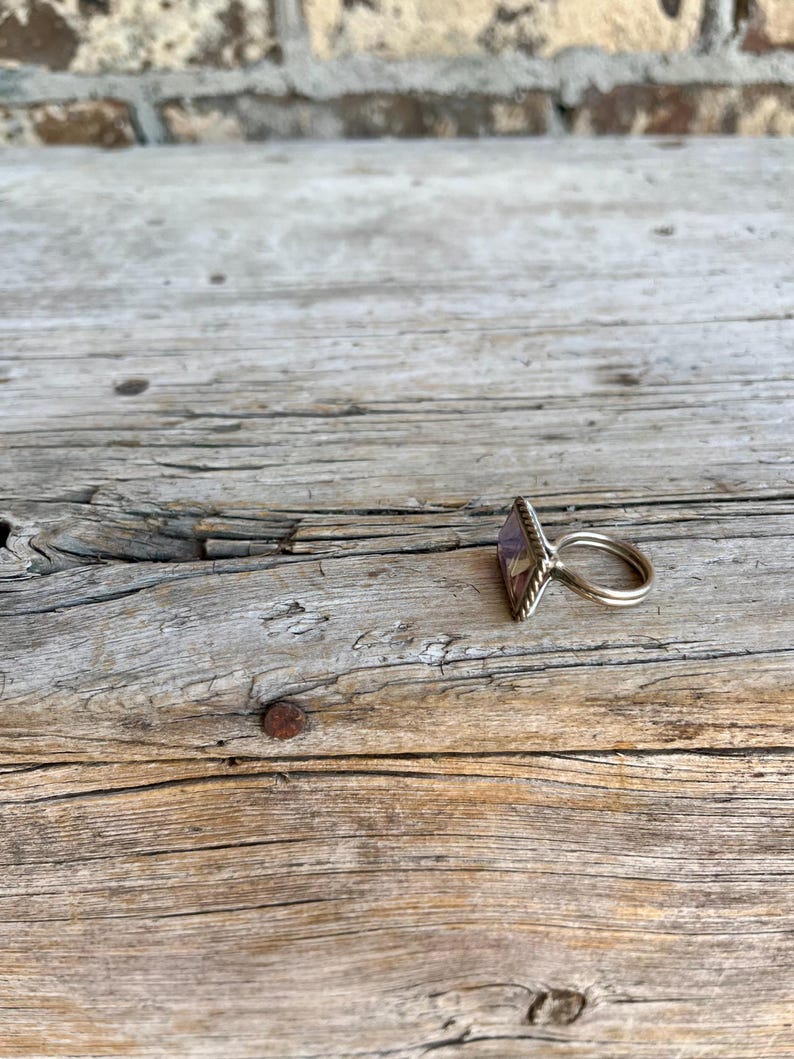 K&ouml;nnte beinhalten: Ein Silberring mit einem rechteckigen, lila Edelstein. Der Ring liegt auf einer rustikalen Holzoberfl&auml;che, die sein Design hervorhebt. Der Edelstein hat einen Facettenschliff und der Ring hat einen einfachen Ring.
