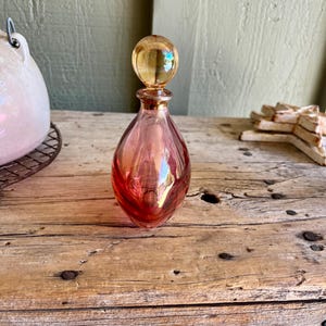 Puede incluir: Un frasco de perfume de vidrio vintage en forma de l&aacute;grima. La botella es un degradado de rosa y rojo con un tap&oacute;n esf&eacute;rico de color &aacute;mbar. La botella se encuentra sobre una superficie de madera envejecida, mostrando su elegante dise&ntilde;o y encanto antiguo.