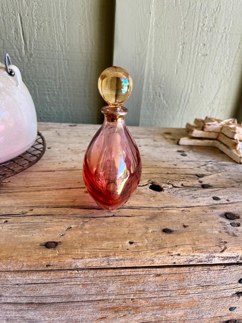 Puede incluir: Un frasco de perfume de vidrio vintage en forma de l&aacute;grima. La botella es un degradado de rosa y rojo con un tap&oacute;n esf&eacute;rico de color &aacute;mbar. La botella se encuentra sobre una superficie de madera envejecida, mostrando su elegante dise&ntilde;o y encanto antiguo.