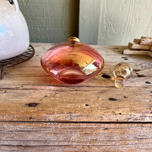 Puede incluir: Un frasco de perfume vintage de vidrio iridiscente con su tap&oacute;n a juego. El frasco tiene forma redondeada con una parte superior dorada y un tono rosa y naranja. El tap&oacute;n es transparente con una parte superior dorada. El frasco est&aacute; sobre una superficie de madera.
