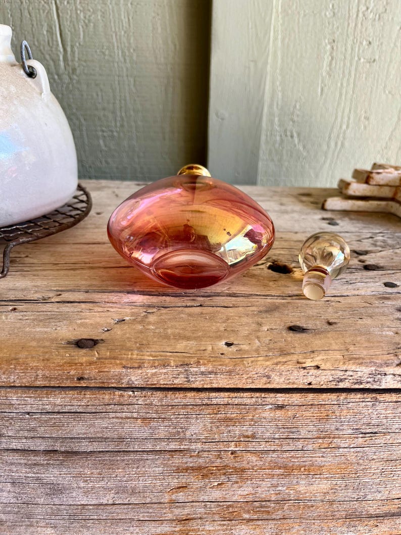 Puede incluir: Un frasco de perfume vintage de vidrio iridiscente con su tap&oacute;n a juego. El frasco tiene forma redondeada con una parte superior dorada y un tono rosa y naranja. El tap&oacute;n es transparente con una parte superior dorada. El frasco est&aacute; sobre una superficie de madera.