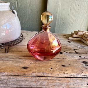 Puede incluir: Un frasco de perfume de vidrio rosa con un cuerpo redondo y un tap&oacute;n esf&eacute;rico. El frasco tiene un cuello y un tap&oacute;n de color dorado. El frasco est&aacute; sobre una superficie de madera.