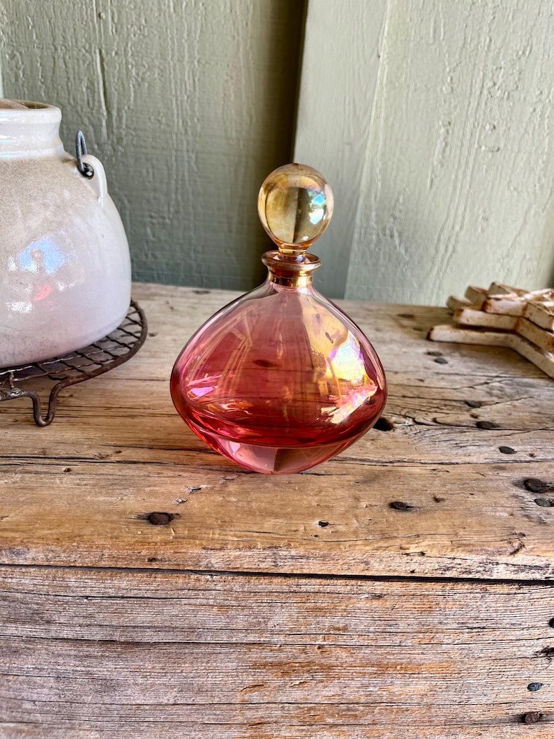 Puede incluir: Un frasco de perfume de vidrio rosa con un cuerpo redondo y un tap&oacute;n esf&eacute;rico. El frasco tiene un cuello y un tap&oacute;n de color dorado. El frasco est&aacute; sobre una superficie de madera.