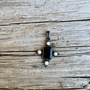 May include: A silver pendant with a rectangular, dark red gemstone at its center, surrounded by four small, round, white pearls. The pendant is in the shape of a cross. The background is a weathered, wooden surface.