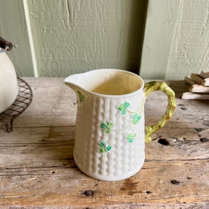 May include: A white ceramic creamer with a green shamrock pattern and a green handle. The creamer has a woven pattern and is in the shape of a pitcher.