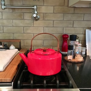 May include: A bright red enamel tea kettle with a wooden handle sits on a black gas stovetop. The kettle is on a burner with a flame underneath. The stovetop is in a kitchen with a tiled backsplash.