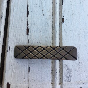 May include: A rectangular, bronze-colored cabinet pull with a diamond pattern.
