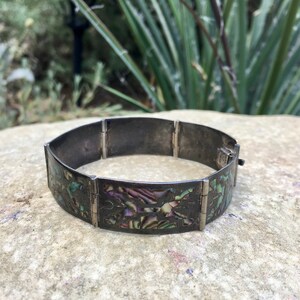 Peut inclure: Un bracelet en argent avec un design à charnières et des accents en nacre d'ormeau incrustés. La nacre est irisée avec des nuances de vert, de bleu et de violet.