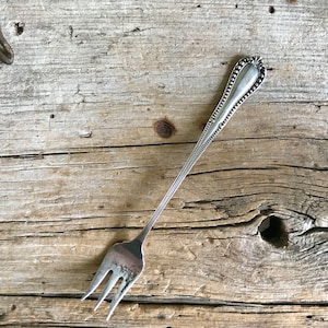 May include: A silver fork with a beaded handle and a simple design. The fork is lying on a wooden surface.