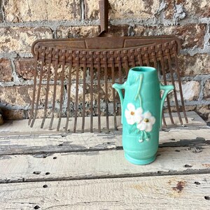 May include: A vintage turquoise vase with white floral accents and handles, set against a weathered wooden surface. A rusty metal garden rake is in the background, with a brick wall behind it.