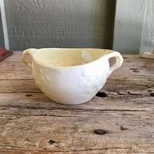May include: A small, white ceramic creamer with two handles. The creamer has a delicate floral design and a slightly yellowed hue.