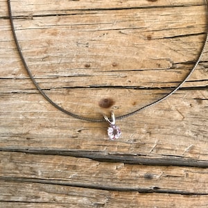 May include: A delicate necklace featuring a small, round, light purple gemstone pendant. The pendant is set in a silver-colored metal and hangs from a thin, dark gray chain. The necklace rests on a weathered, light brown wooden surface.