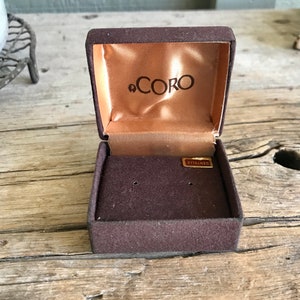 May include: A brown velvet jewelry box with a gold label that reads "Coro" on the inside of the lid. The box has a brown velvet lining and two small holes for earrings.