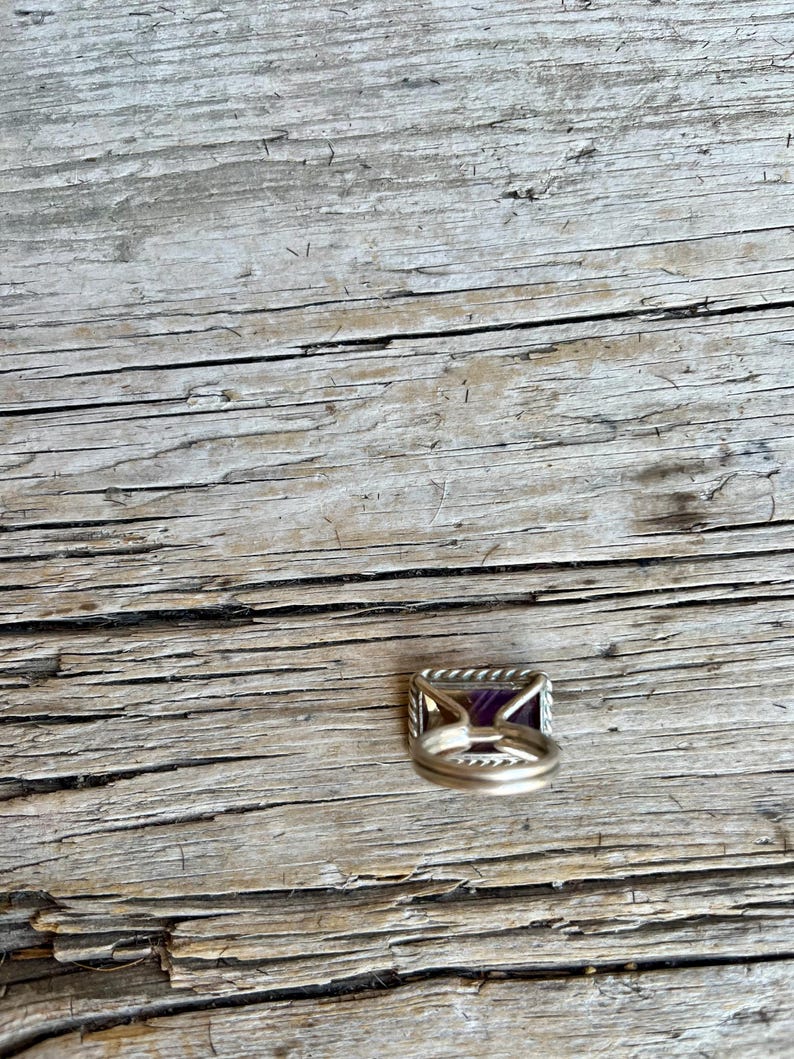 K&ouml;nnte beinhalten: Ein silberner Ring mit einem rechteckigen, tiefvioletten Edelstein. Der Ring hat ein gedrehtes Seildesign um den Edelstein und ein einfaches Band. Der Ring liegt auf einer verwitterten Holzoberfl&auml;che.
