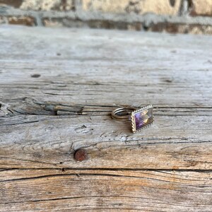 K&ouml;nnte beinhalten: Ein Silberring mit einem rechteckigen, mehrfarbigen Edelstein. Der Edelstein hat Schattierungen von Lila und Gelb. Der Ring hat ein geflochtenes Design um den Edelstein.