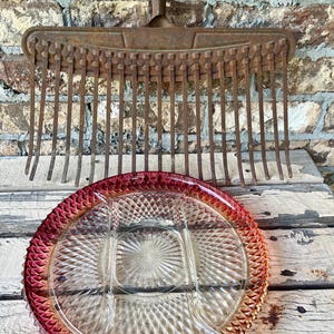 May include: A vintage metal garden rake with a long handle and multiple tines positioned behind a round, clear glass divided serving tray featuring a textured diamond pattern and a deep red gradient rim around the outer edge of the dish.