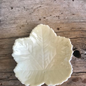 May include: A white ceramic leaf-shaped dish with a raised design. The dish is on a wooden surface.