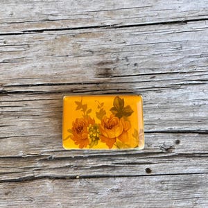 May include: Rectangular, yellow decorative tile with a floral design featuring orange roses and green leaves. The tile is set against a weathered, wooden background, showcasing its vintage aesthetic.