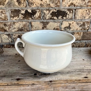 May include: A white ceramic bowl with a handle. The bowl has a rounded shape and a wide rim. The background is a weathered brick wall and a wooden surface.