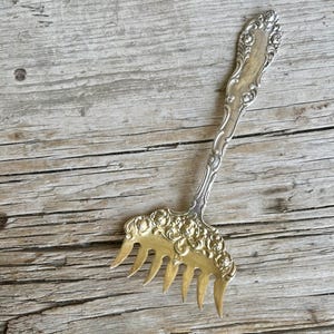 May include: Silver-plated spaghetti server with a decorative handle and a gold-toned tines. The server has an ornate floral design on the handle and the tines. The server is placed on a weathered wooden surface.