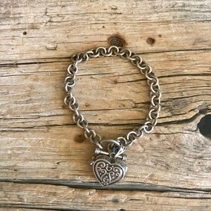 May include: A silver chain bracelet with a heart-shaped charm. The charm has an intricate design and is attached to the chain with a small ring.