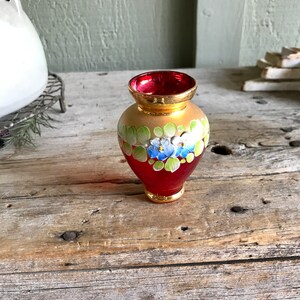 May include: A small, red glass vase with gold accents and hand-painted blue and white flowers. The vase has a rounded body and a slightly flared opening.