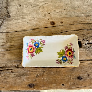 May include: A rectangular, white porcelain dish with a gold rim and floral designs. The flowers are in shades of red, yellow, blue, and purple. The dish is placed on a weathered wooden surface.