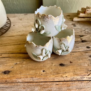 May include: Three white porcelain egg cups with a gold rim and a delicate floral design of lily of the valley.