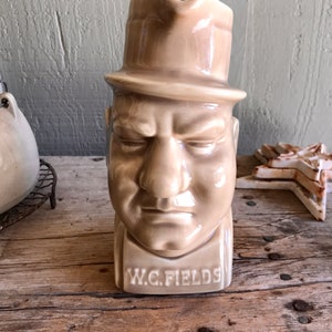 May include: A ceramic pitcher shaped like the head of a man wearing a hat. The pitcher is beige and has the words "W.C. FIELDS" written on the front.