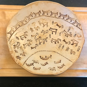 May include: A ceramic plate with a brown glaze and a design of stylized animals and boats. The design is in a dark brown color and is reminiscent of ancient petroglyphs.