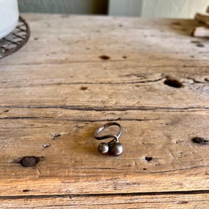 May include: A silver ring with two round balls attached to the band. The ring is on a wooden surface.