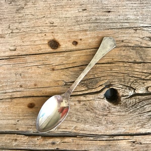 May include: A silver teaspoon with a rounded bowl and a simple handle sits on a weathered wooden surface.